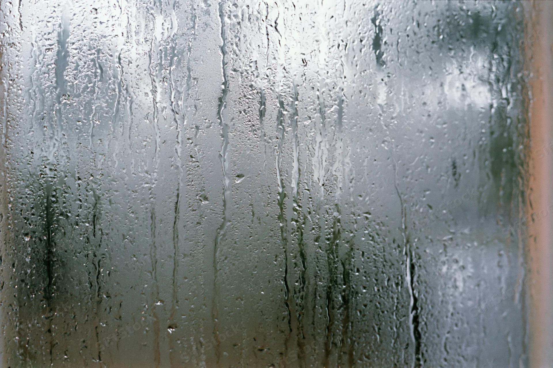 Fogged window glass covered in water droplets and streaks from condensation, showing blurred outdoor view through translucent surface