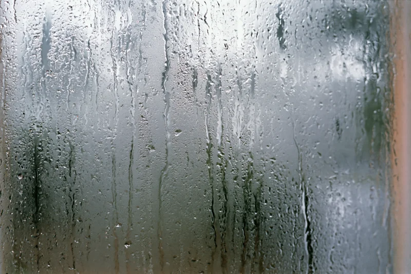 Fogged window glass covered in water droplets and streaks from condensation, showing blurred outdoor view through translucent surface