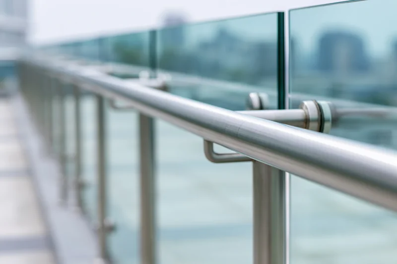 Close-up of a modern glass balcony railing with a brushed stainless steel handrail and vertical glass panels, set against a blurred city view