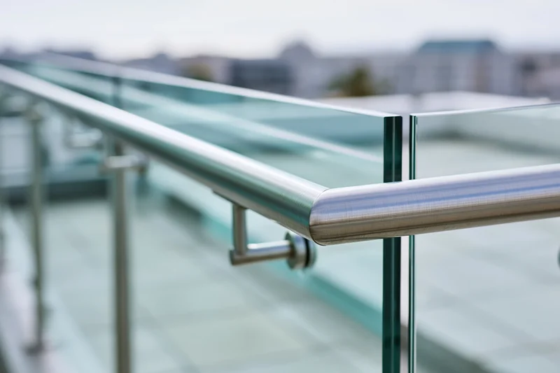 Close-up of a modern glass balcony railing with a brushed stainless steel handrail on an urban building exterior