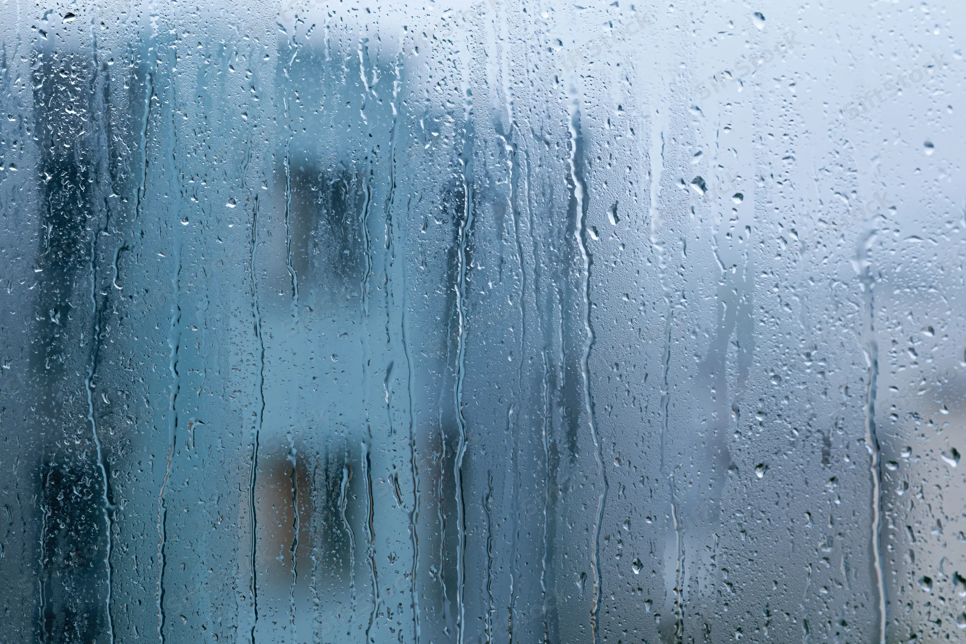 Rain-covered window glass with water droplets and streaks, showing blurred outdoor view through transparent surface