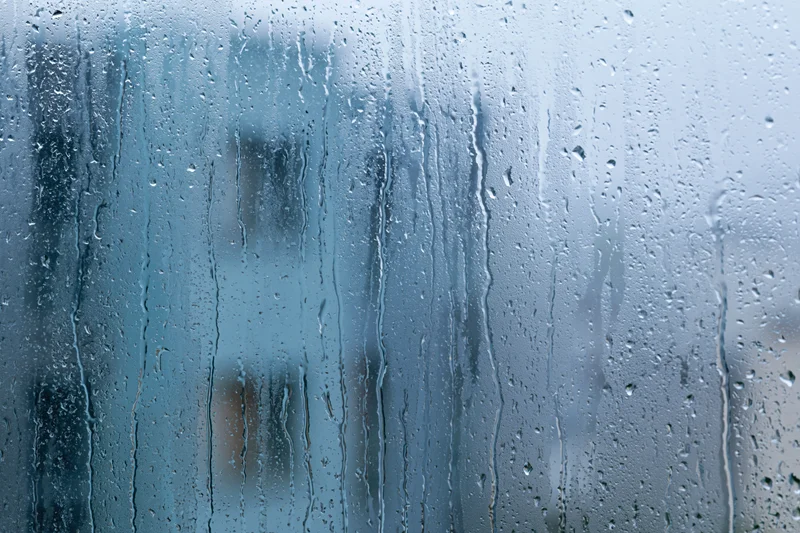 Rain-covered window glass with water droplets and streaks, showing blurred outdoor view through transparent surface