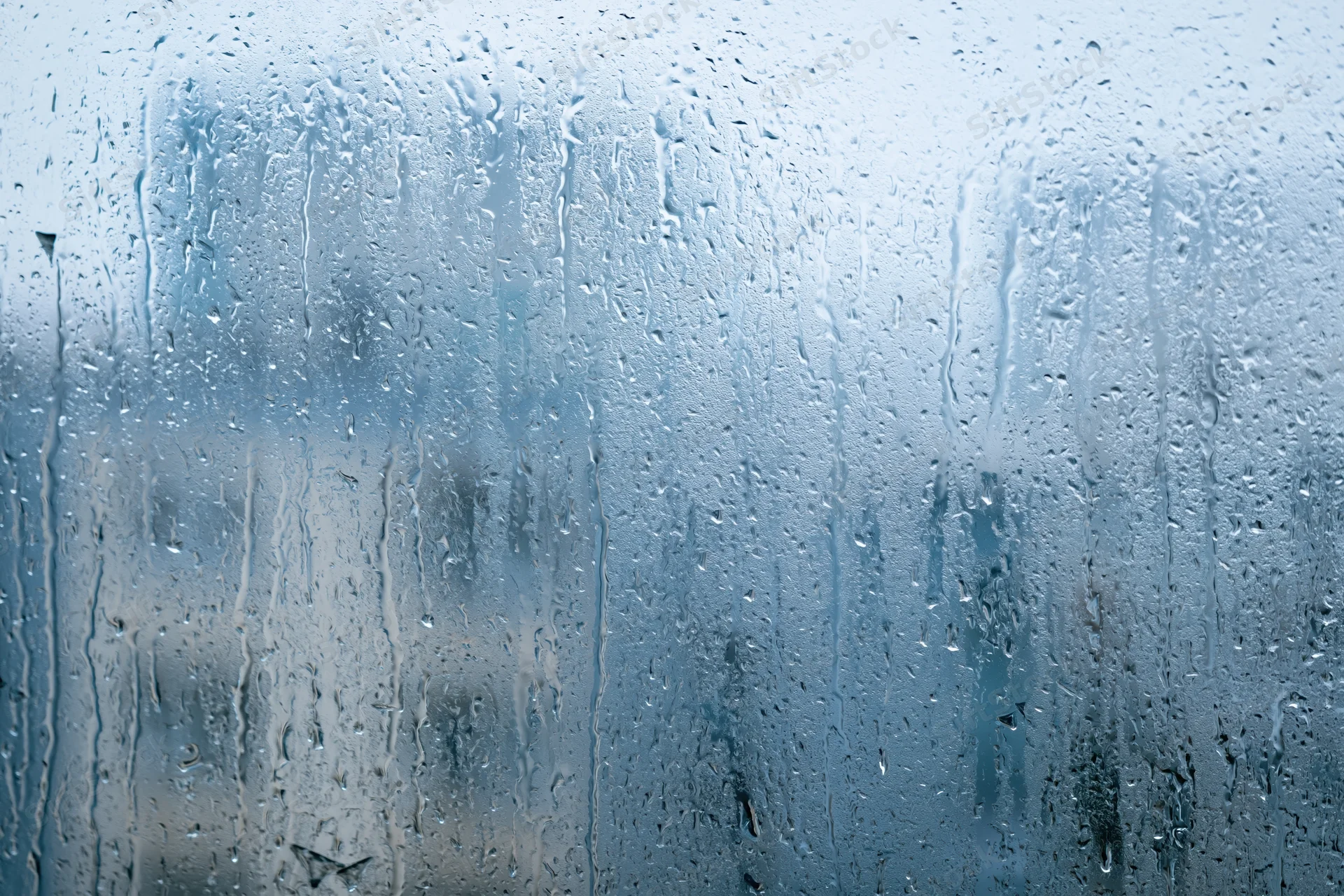 Rain-covered window glass with water droplets and streaks, showing blurred outdoor view through wet surface