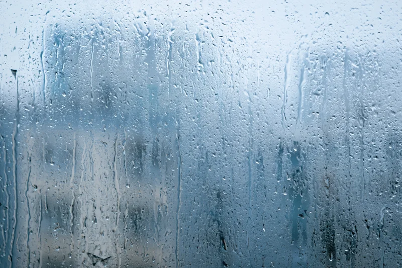Rain-covered window glass with water droplets and streaks, showing blurred outdoor view through wet surface