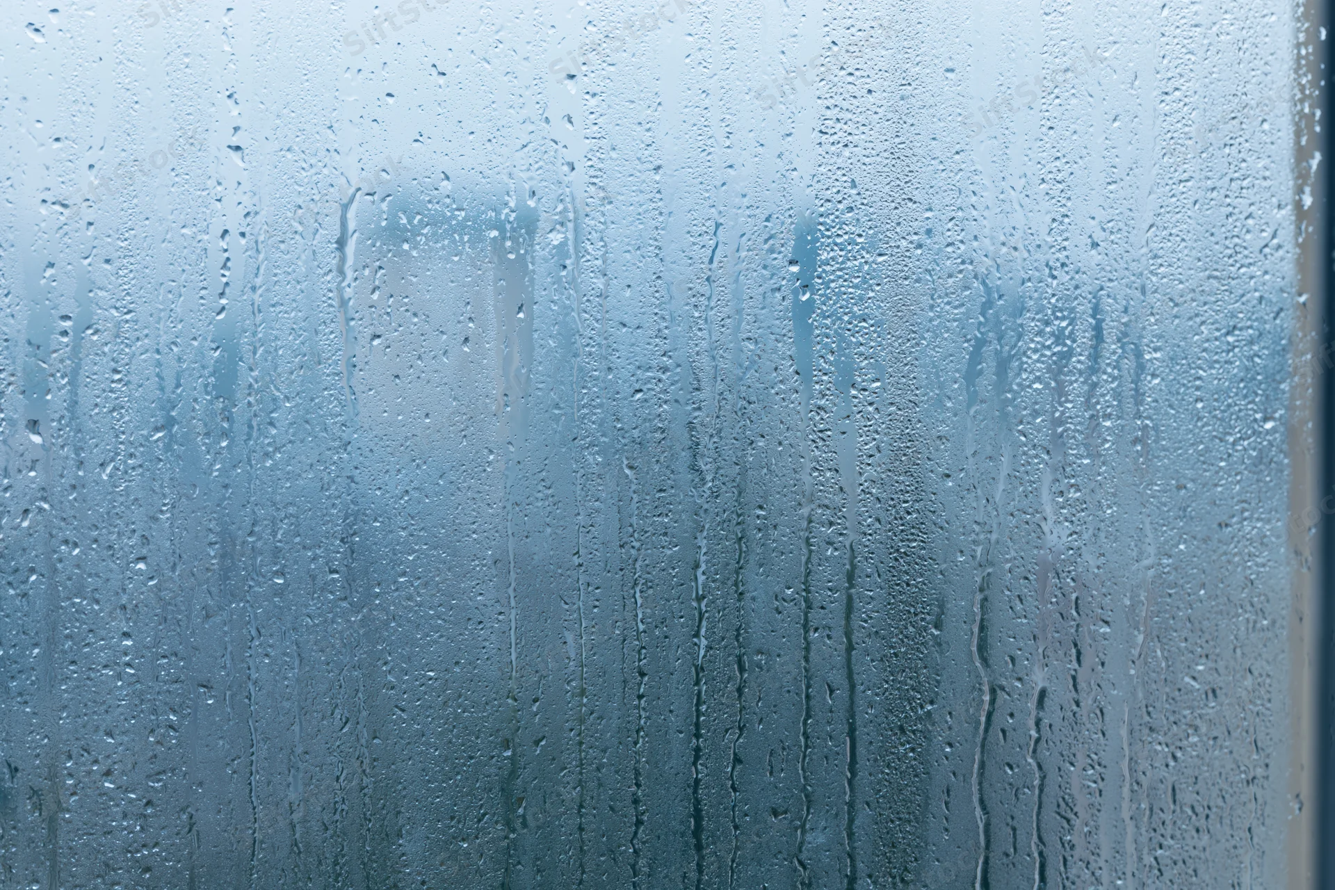 Rain-covered window glass with water droplets and streaks, showing blurred outdoor view through wet surface