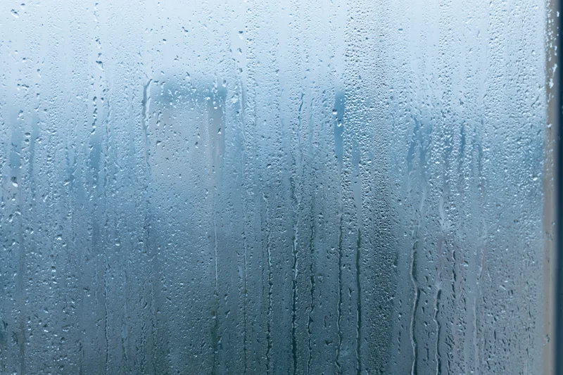 Rain-covered window glass with water droplets and streaks, showing blurred outdoor view through wet surface