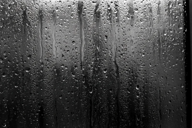 Rain-streaked dark glass window surface covered in water droplets and vertical streaks