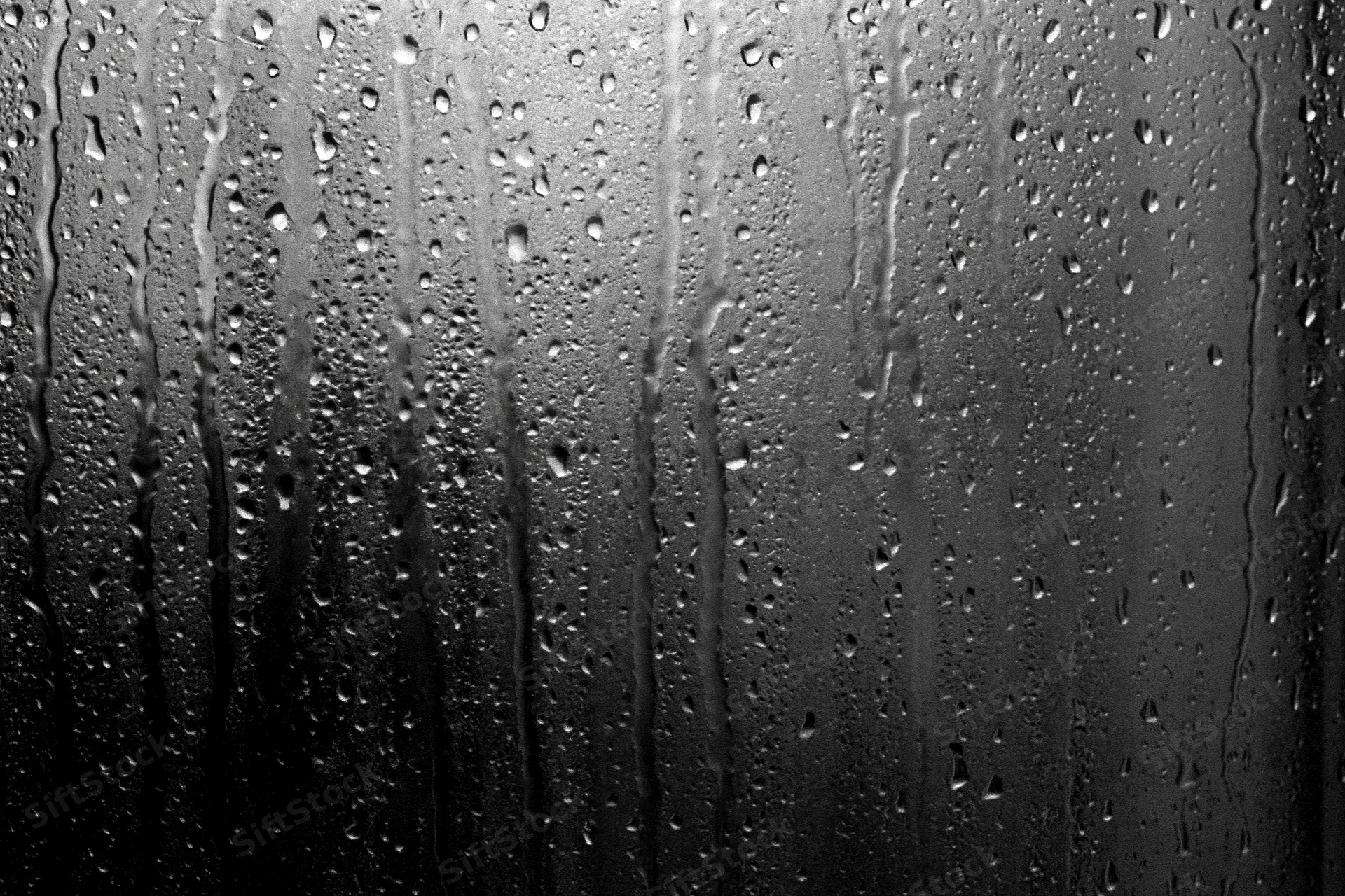 Rain-streaked dark glass window surface covered in water droplets and vertical streaks