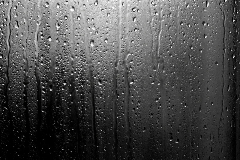 Rain-streaked dark glass window surface covered in water droplets and vertical streaks
