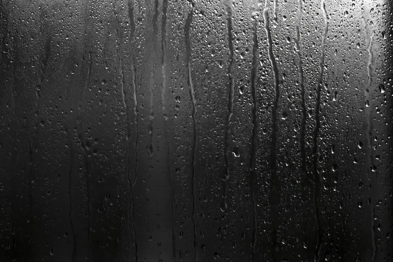 Rain-streaked dark glass window surface with water droplets and vertical streaks