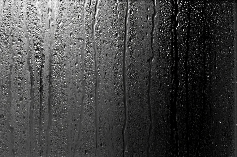 Rain-streaked dark glass window surface covered in water droplets and vertical streaks