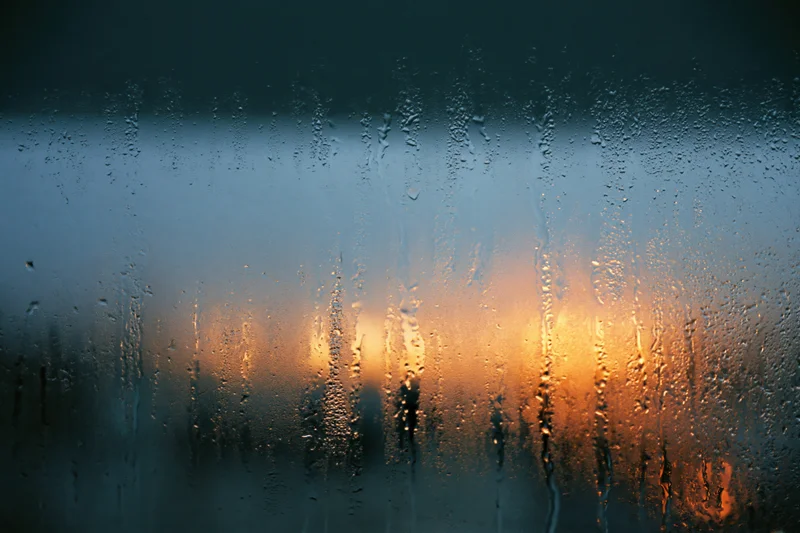 Rain-streaked window glass with condensation droplets and blurred city lights at twilight