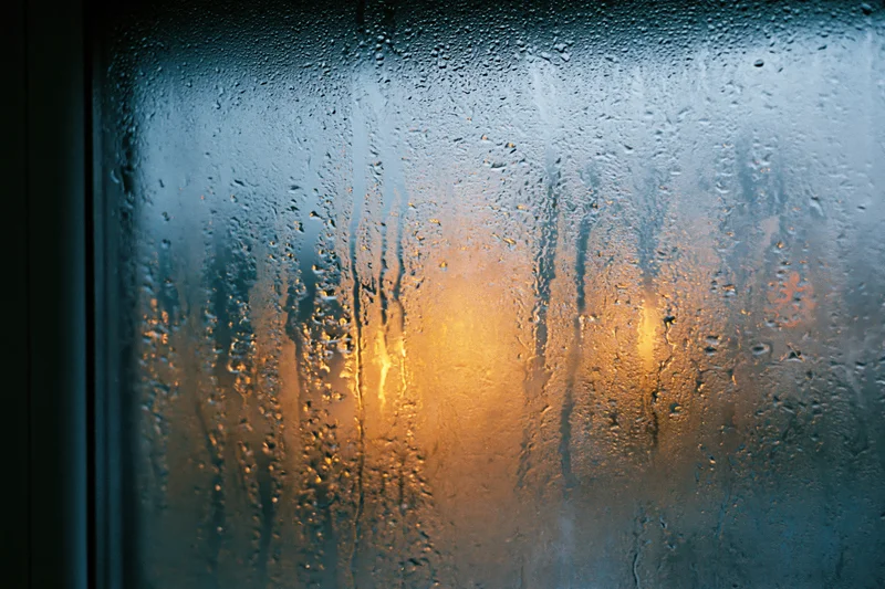 Rain-streaked window glass with condensation droplets and warm light reflection at night