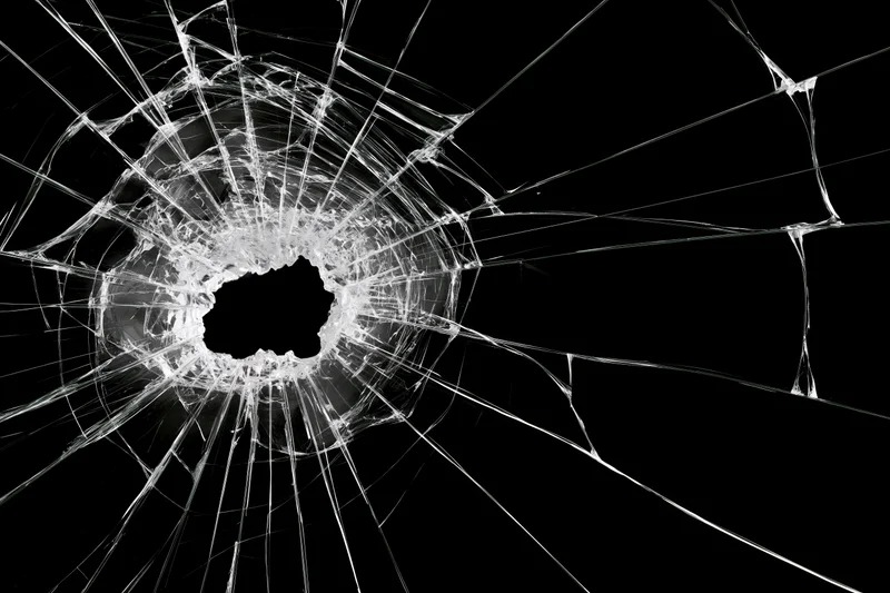 Shattered glass pane with central impact hole and radiating cracks against black background