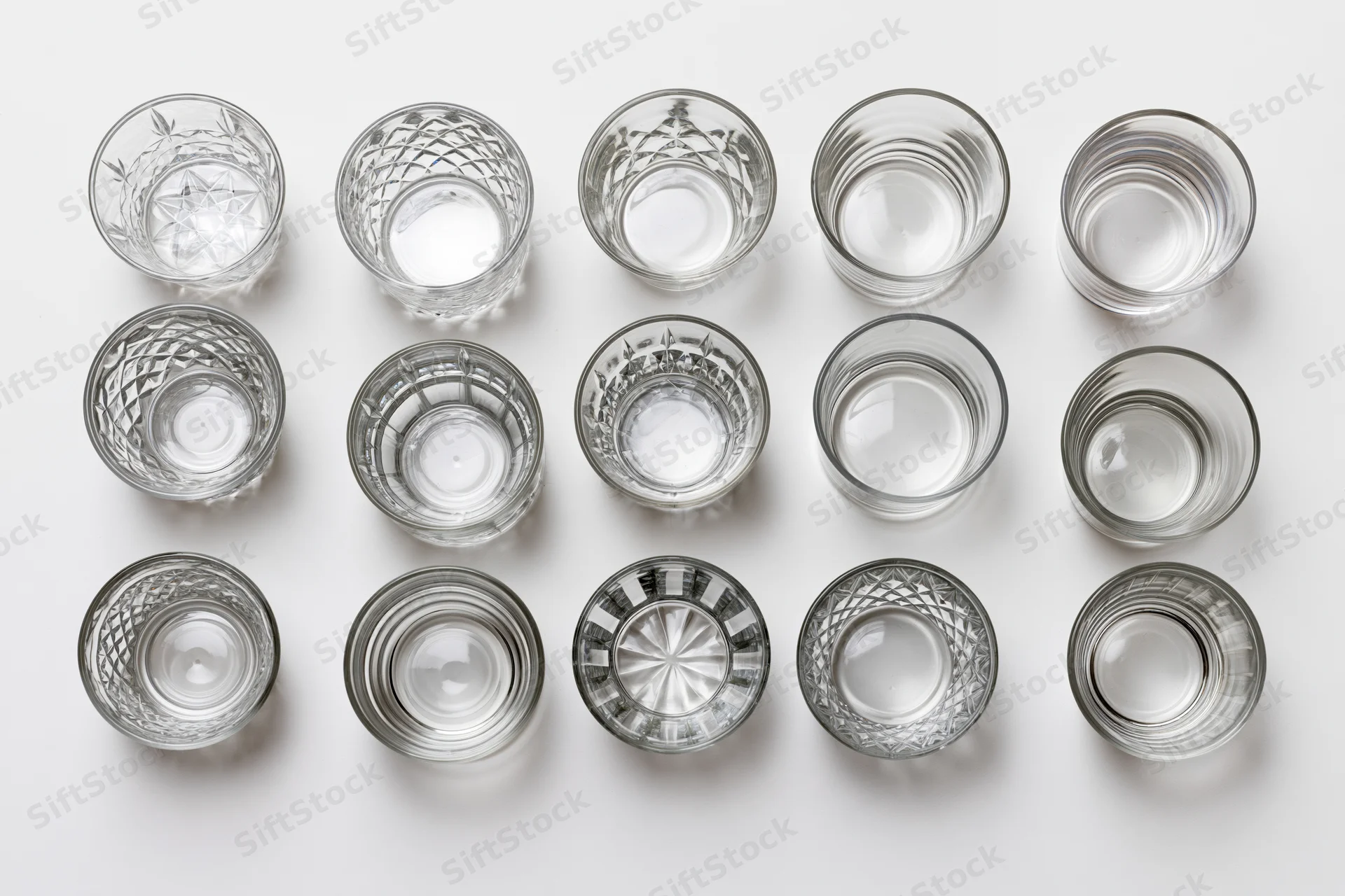 Eighteen empty whiskey tumblers and rocks glasses arranged in a grid on a white surface