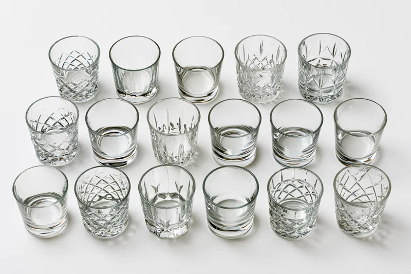 Eighteen empty whiskey tumblers and rocks glasses arranged in rows on a white background