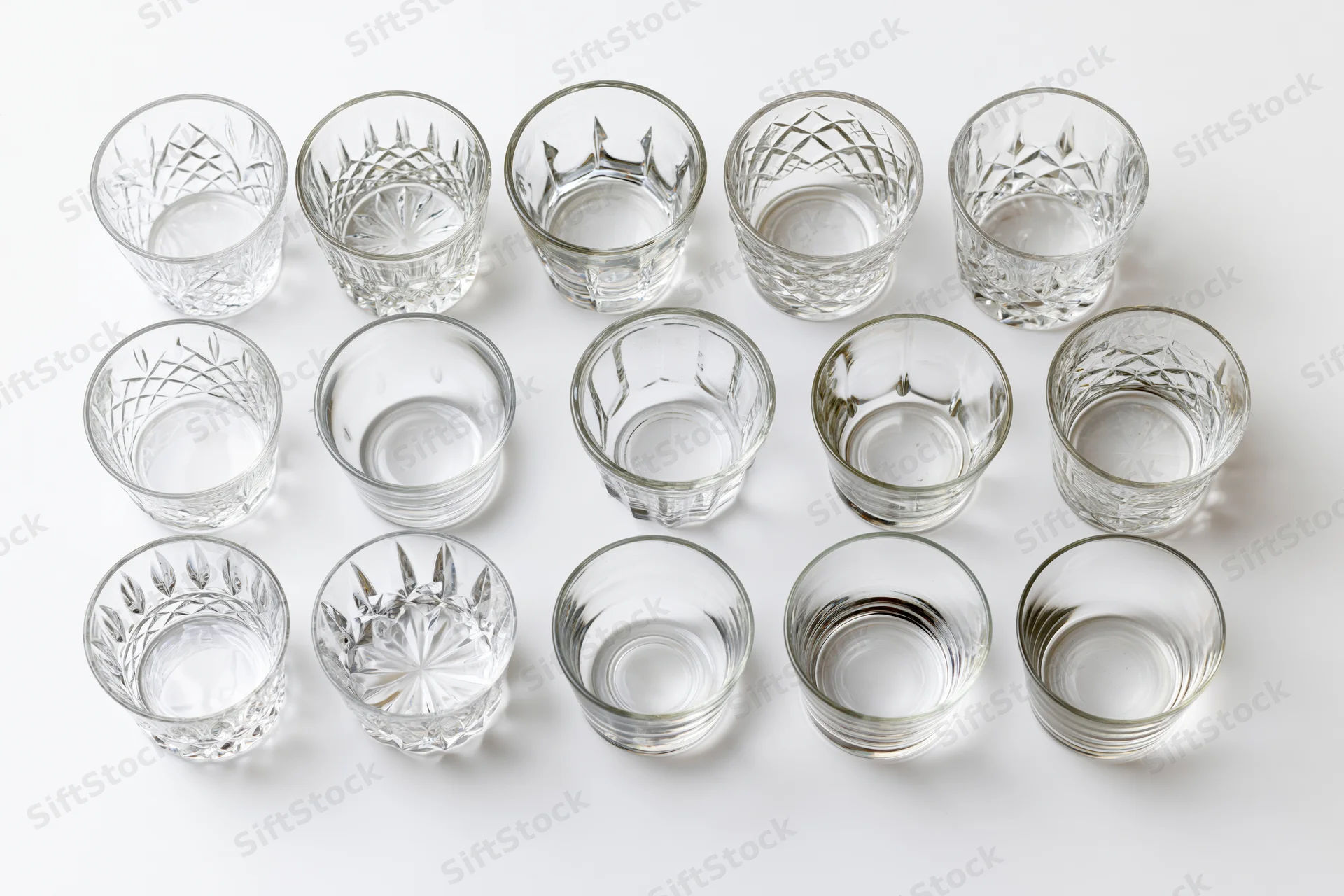 Eighteen empty whiskey tumblers and rocks glasses arranged in three rows on a white background