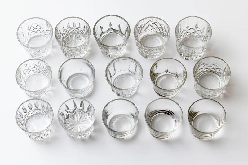 Eighteen empty whiskey tumblers and rocks glasses arranged in three rows on a white background