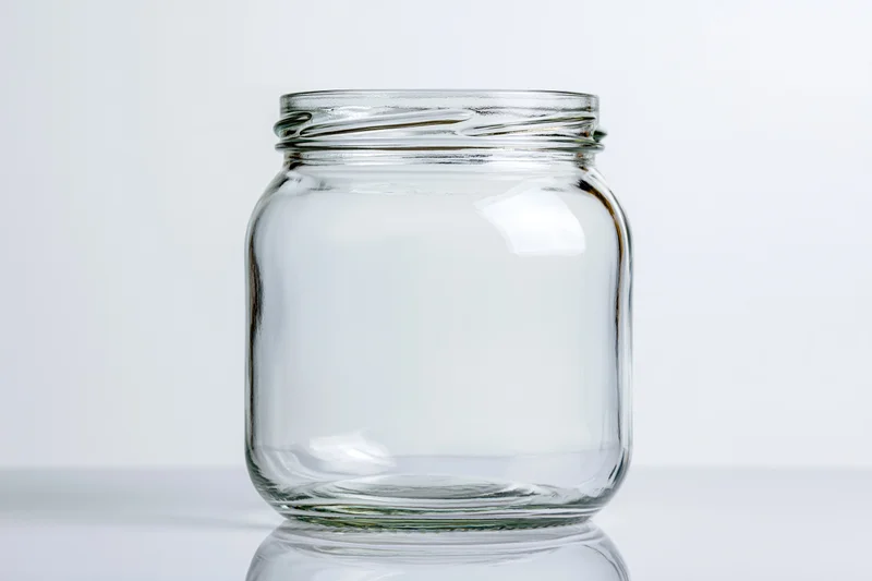 Empty wide-mouth glass jar sitting upright on a white reflective surface
