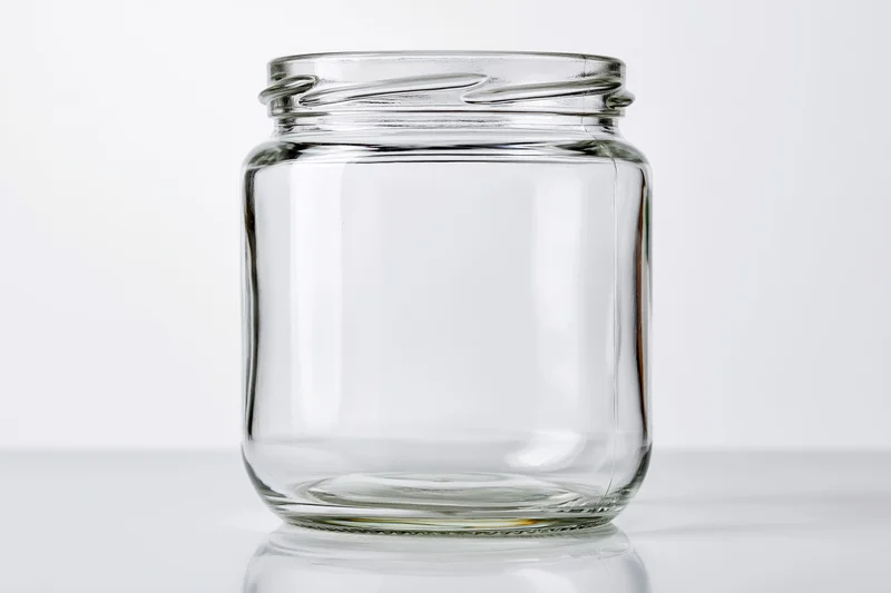 Empty wide-mouth glass jar sitting upright on a white reflective surface