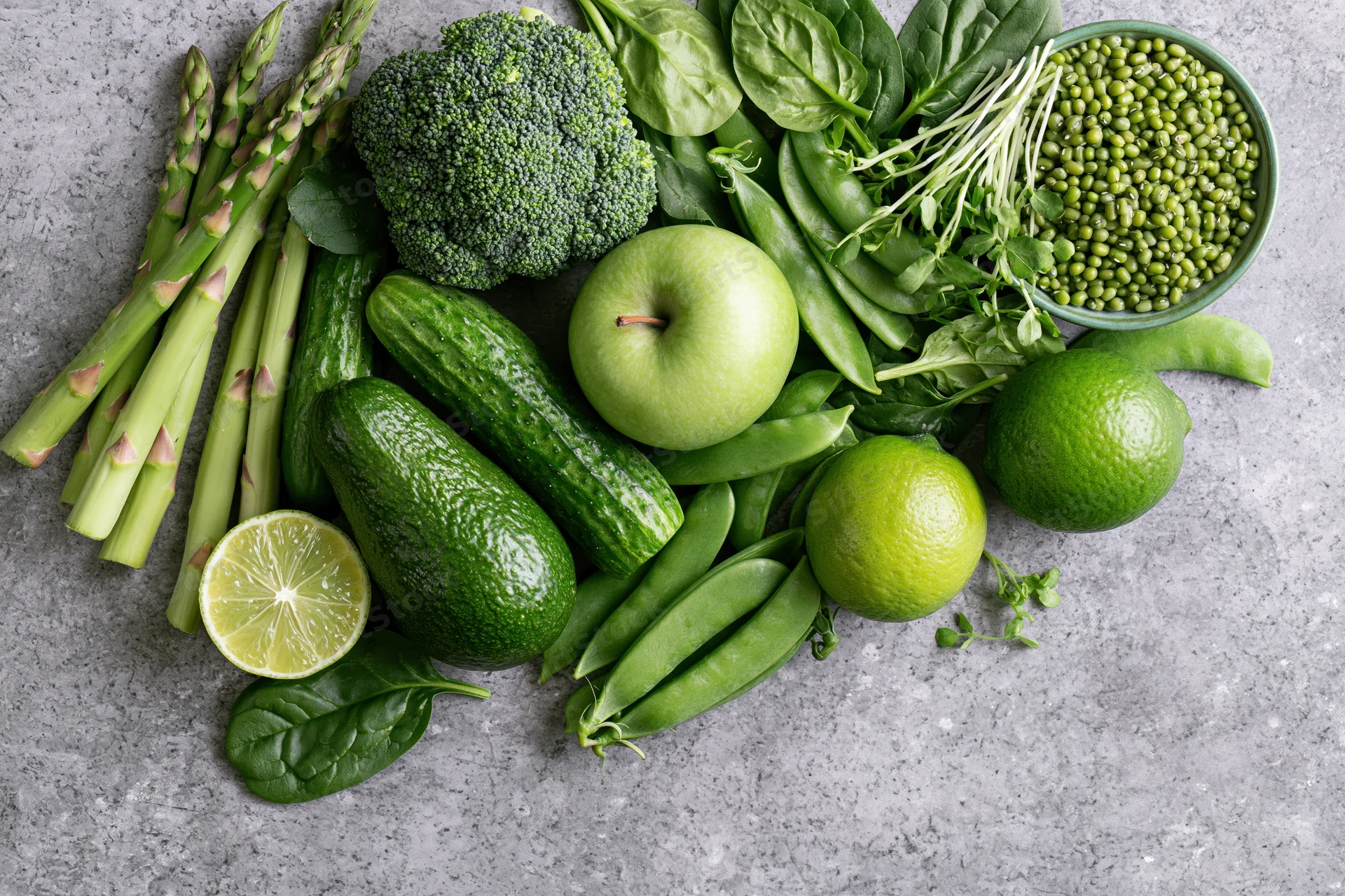 Assorted green vegetables and fruits arranged in a flat lay on a gray stone surface