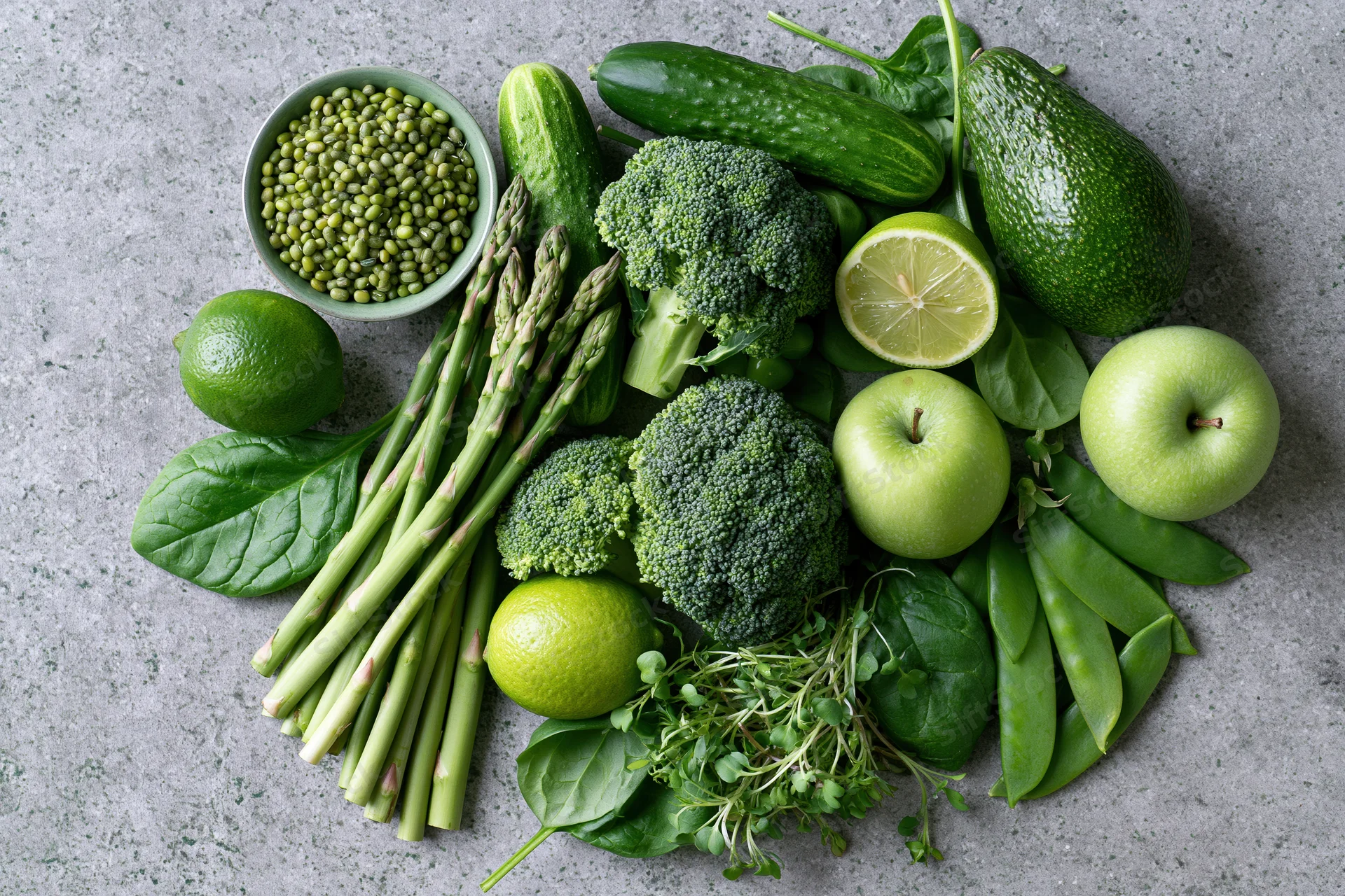 Assorted green vegetables and fruits arranged in a flat lay on a gray surface