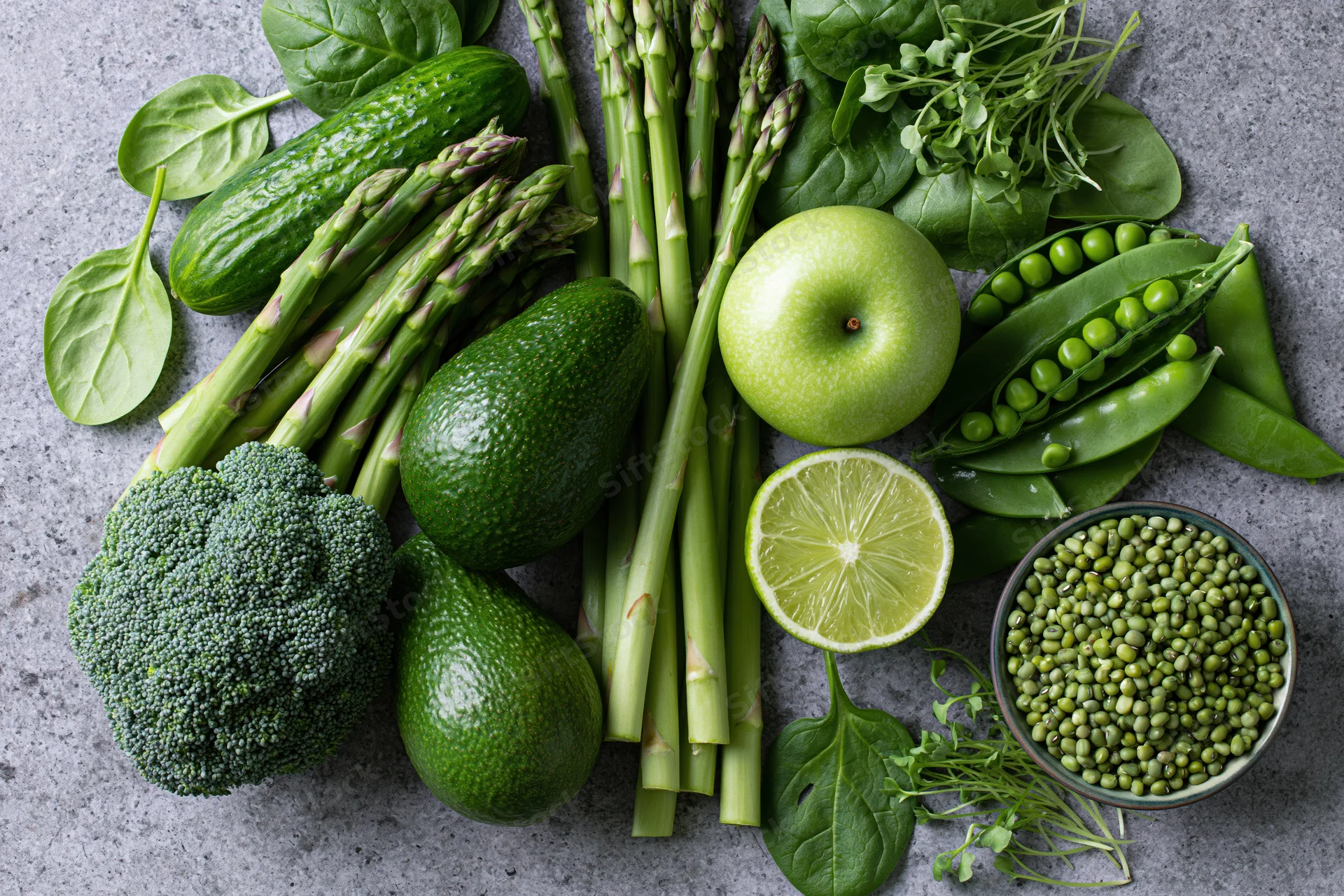 Flat lay of assorted green vegetables and fruits arranged on a gray stone countertop
