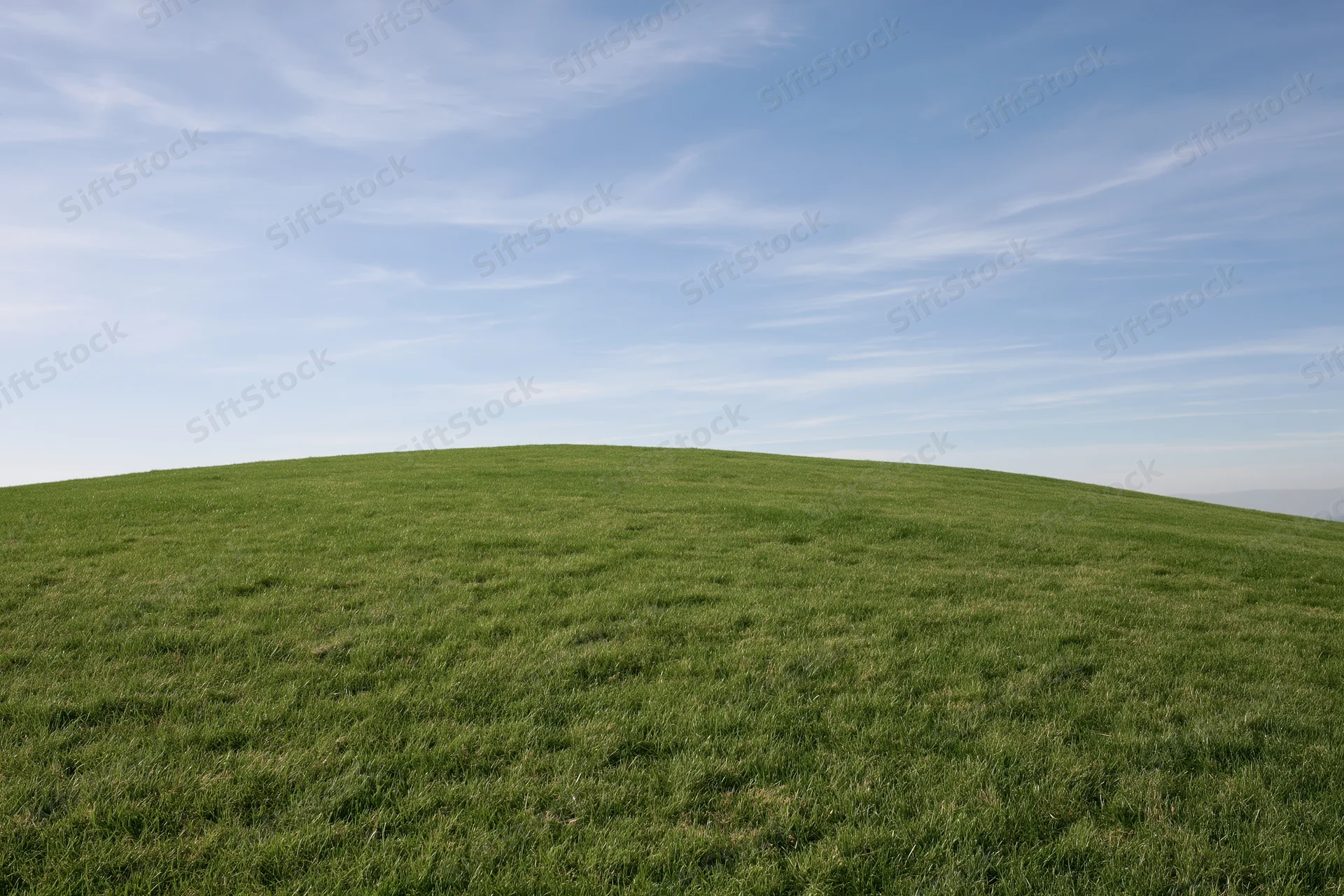 Gently sloping green grass hill stretching to the horizon under a clear blue sky with wispy clouds