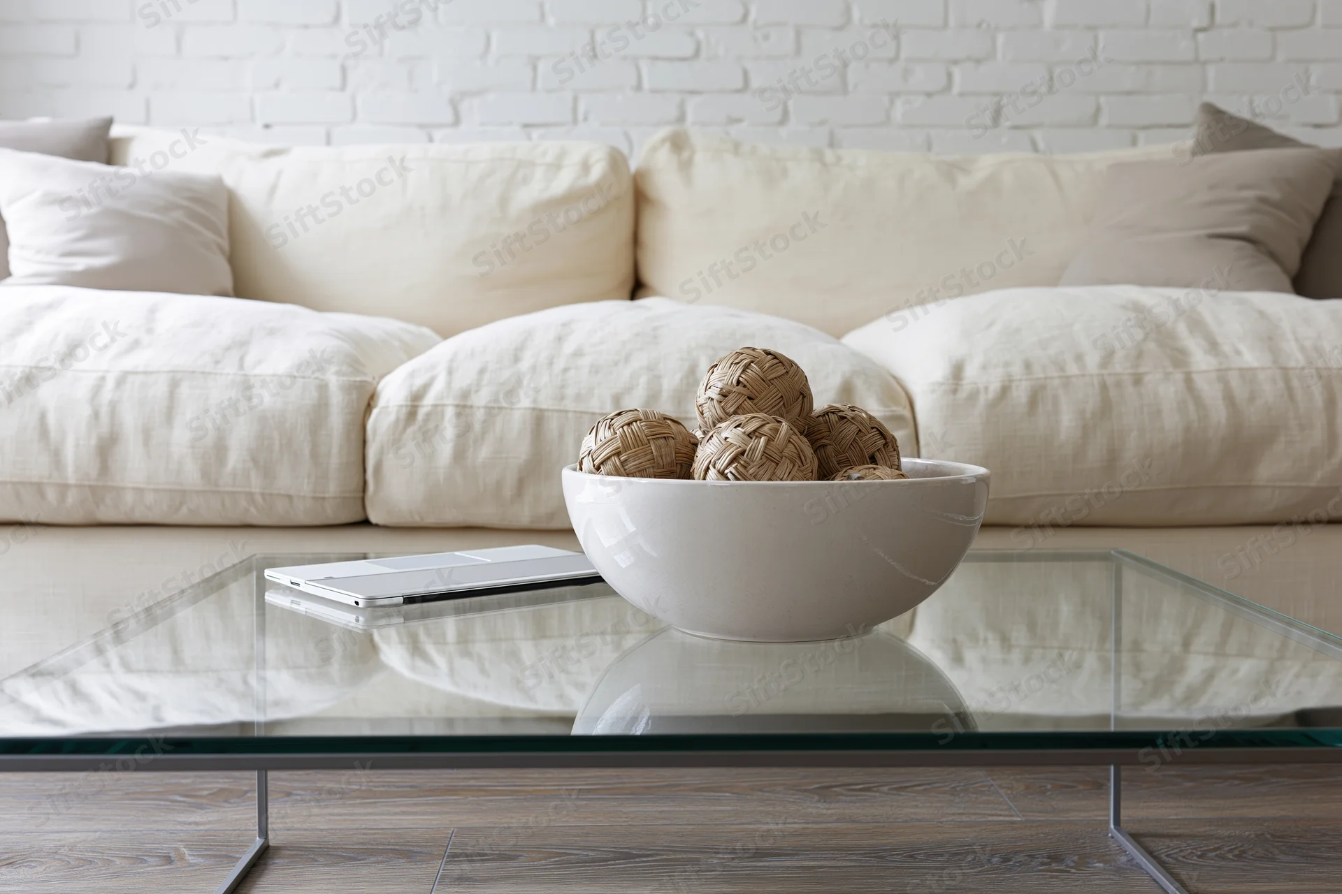 Glass coffee table with thin metal legs holding a white bowl of woven balls and a closed laptop, in front of a cream sofa against a white brick wall