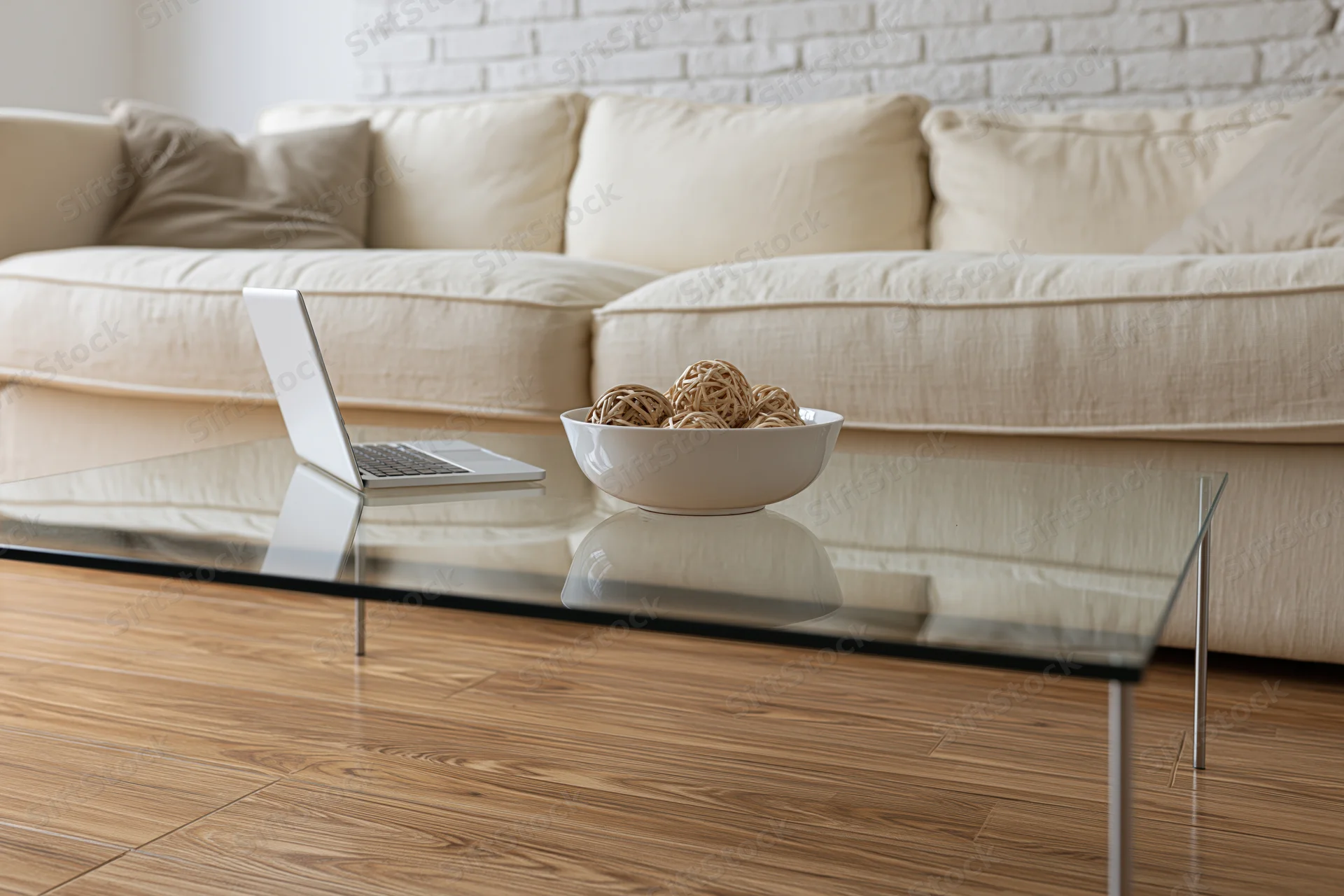 Glass coffee table with thin metal legs holding an open laptop and a white bowl of decorative balls in a living room with a beige sectional sofa and hardwood floor