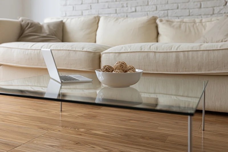 Glass coffee table with thin metal legs holding an open laptop and a white bowl of decorative balls in a living room with a beige sectional sofa and hardwood floor