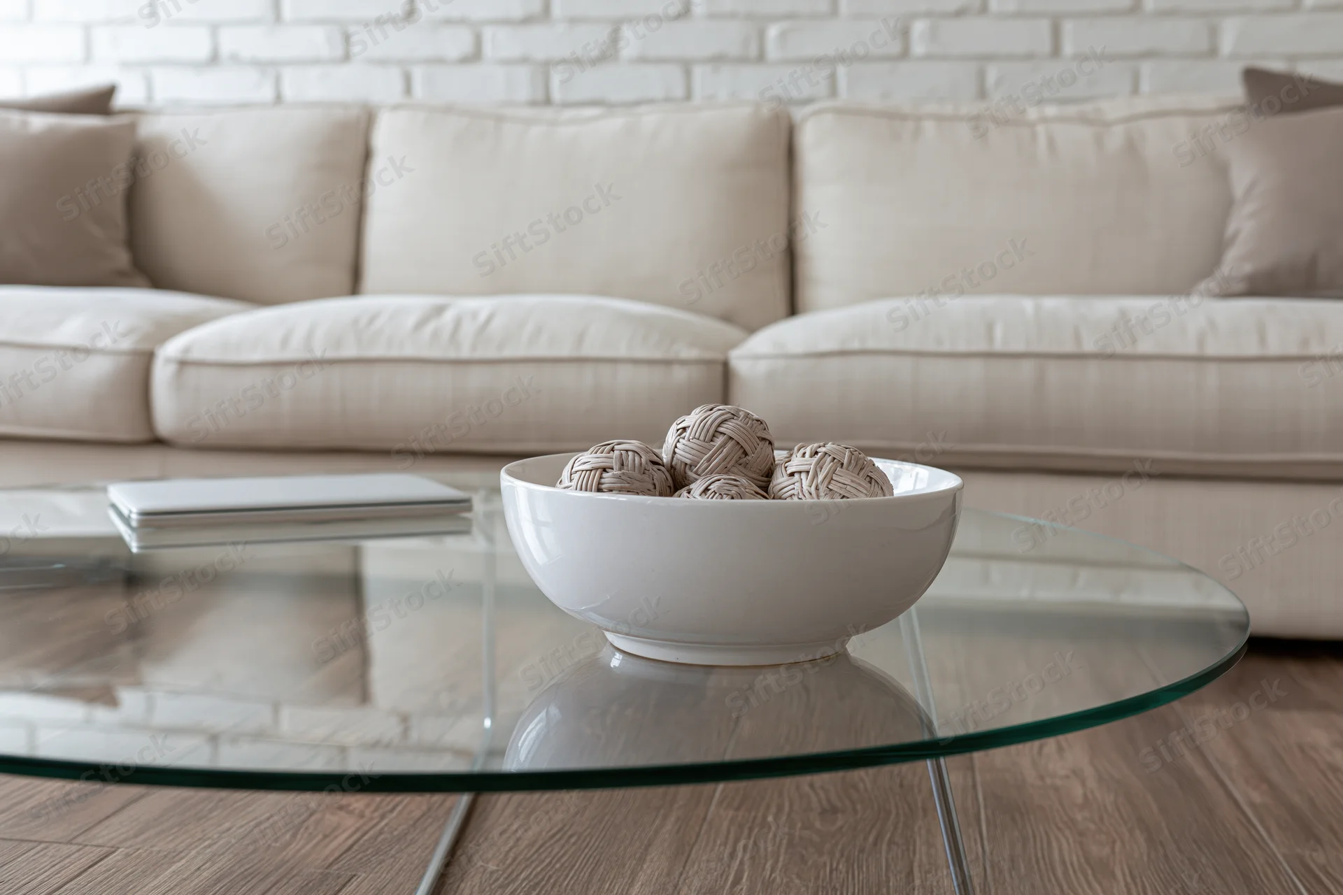 Glass coffee table with thin metal legs holding a white bowl of woven decorative balls in a living room with beige sofa and brick wall