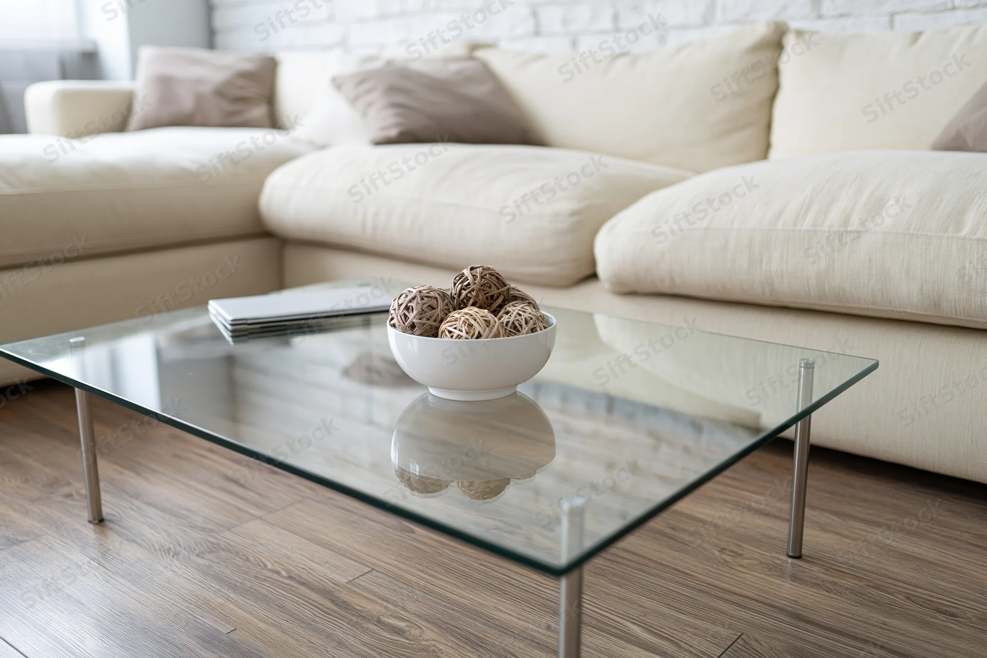 Glass coffee table with thin metal legs holding a white bowl of decorative wicker balls and a stack of magazines in a modern living room with a beige sectional sofa and wood flooring