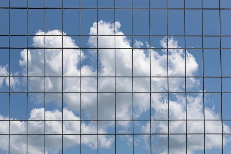 Glass curtain wall facade of a modern office building reflecting blue sky and white clouds
