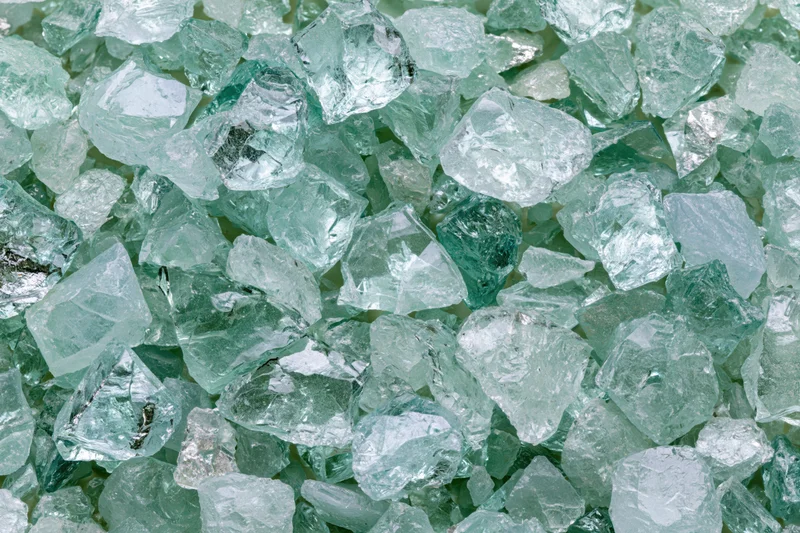 Crushed tempered glass fragments scattered densely in a pile, showing greenish transparent shards with rough edges and reflective surfaces
