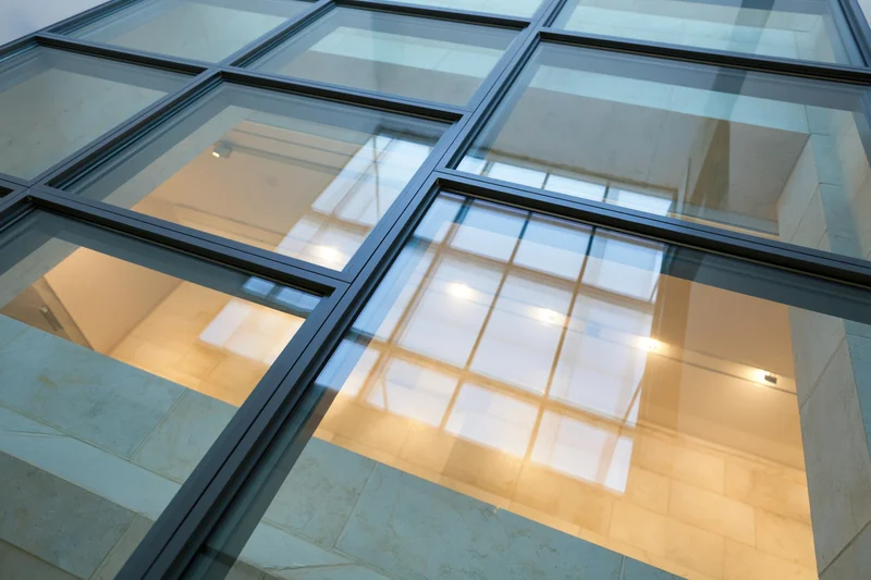 Transparent glass floor panel with black metal grid framing, reflecting warm interior lighting and stone walls