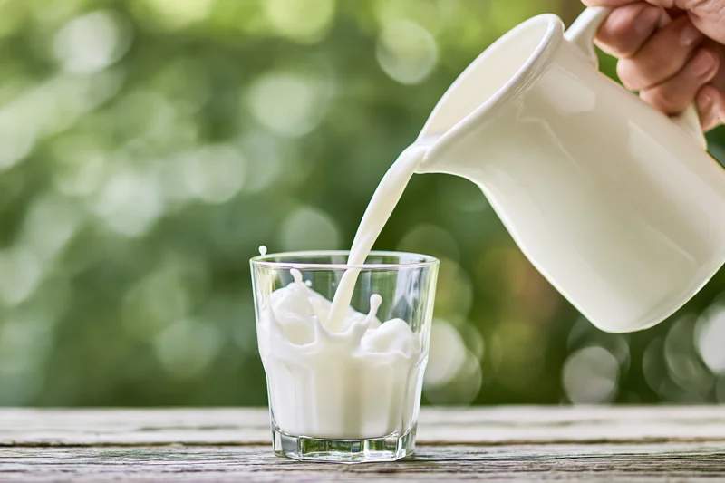 Milk being poured from a white ceramic jug into a clear glass on a wooden table outdoors