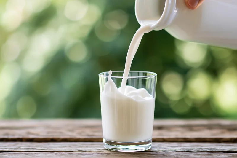 Milk being poured from a white ceramic jug into a clear glass on a wooden table outdoors