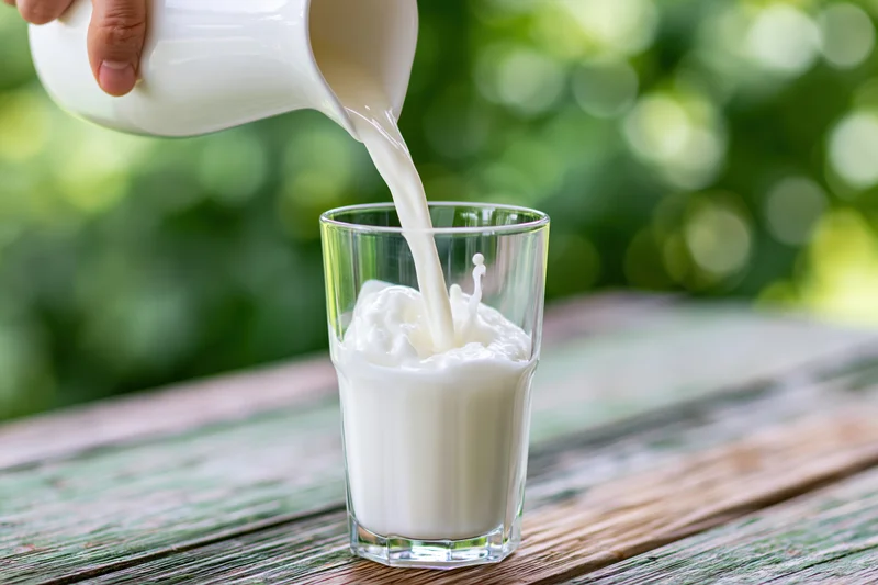 Milk being poured from a white ceramic jug into a clear glass on a wooden table outdoors