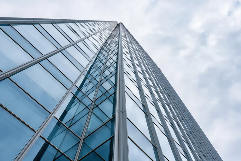 Modern glass curtain-wall skyscraper facade viewed from ground level against cloudy sky