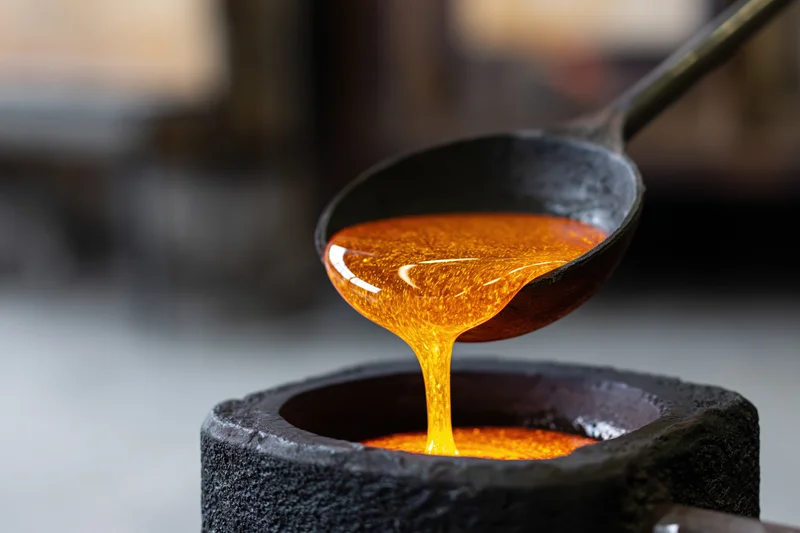 Molten glass pouring from a metal ladle into a dark stone crucible in a workshop setting
