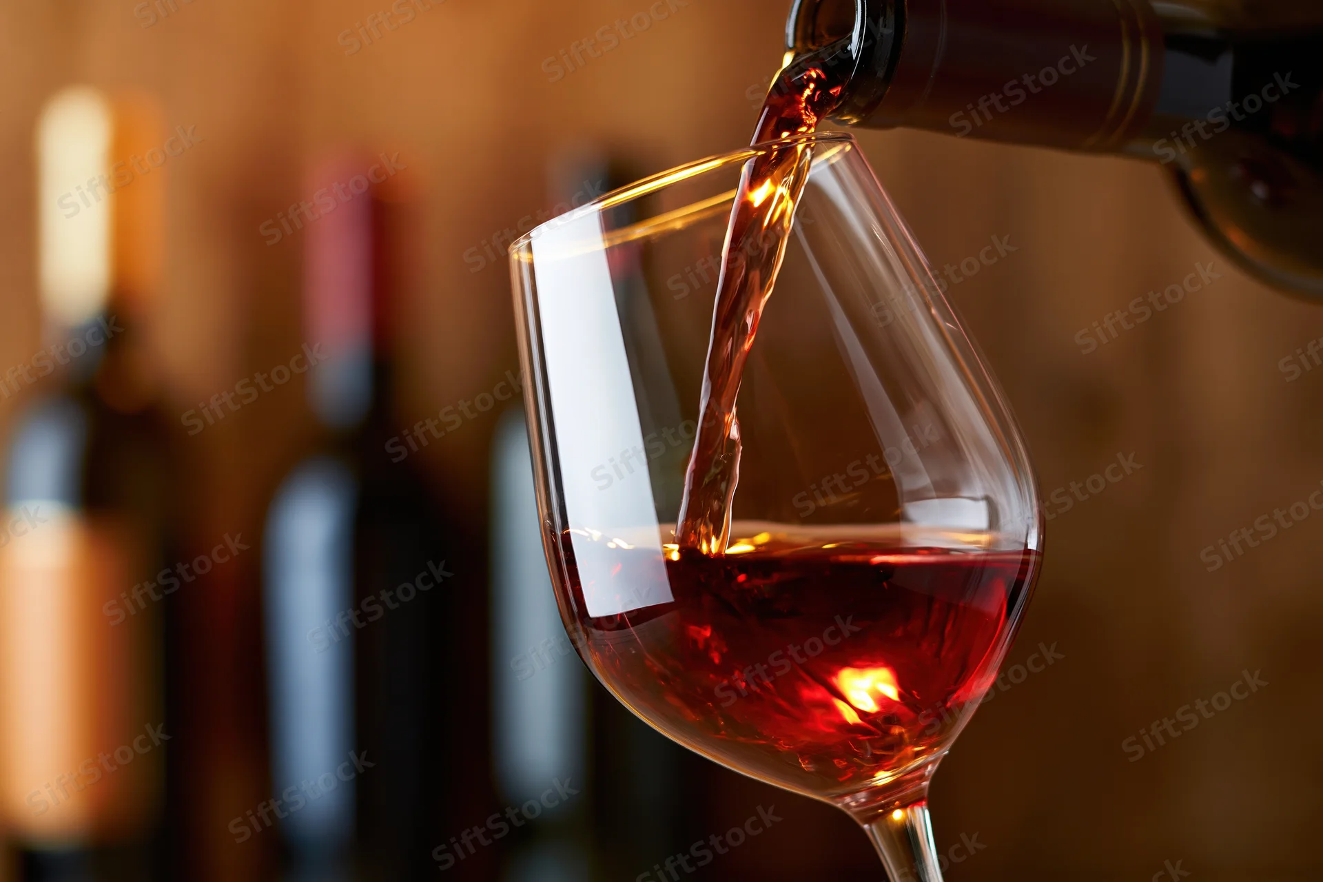 Red wine being poured from a dark bottle into a tilted crystal glass with blurred bottles in the background
