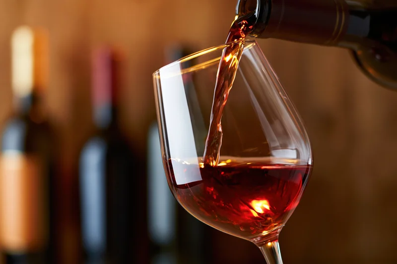 Red wine being poured from a dark bottle into a tilted crystal glass with blurred bottles in the background