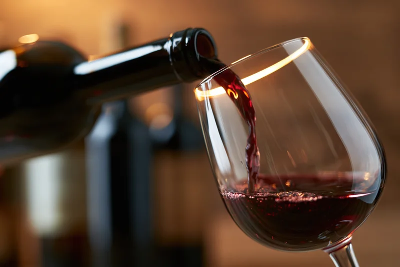 Red wine pouring from a dark bottle into a tilted crystal glass with warm ambient lighting and blurred background