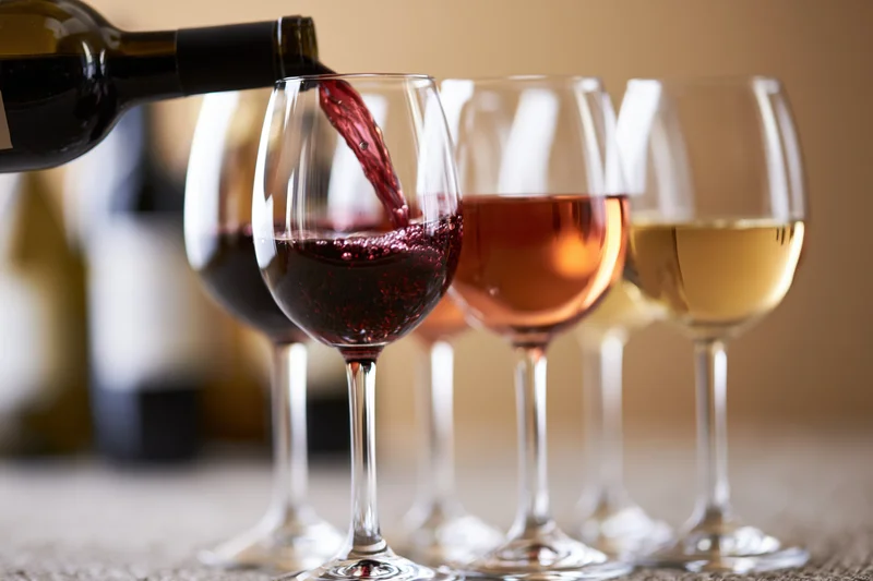 Red wine being poured from a dark bottle into the foremost of four wine glasses arranged in a row on a table