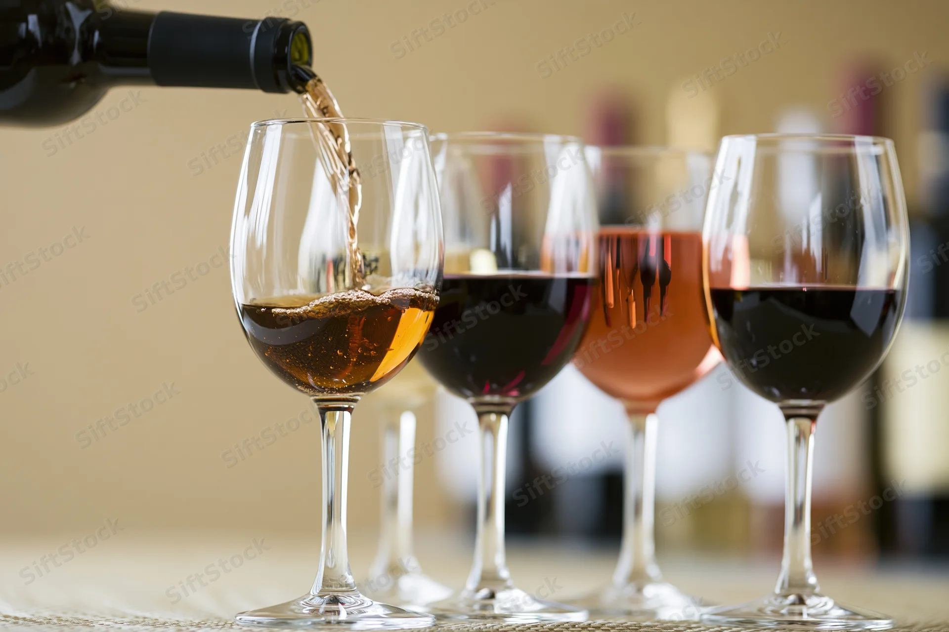 Red wine being poured from a dark bottle into the foremost of four wine glasses arranged in a row on a table