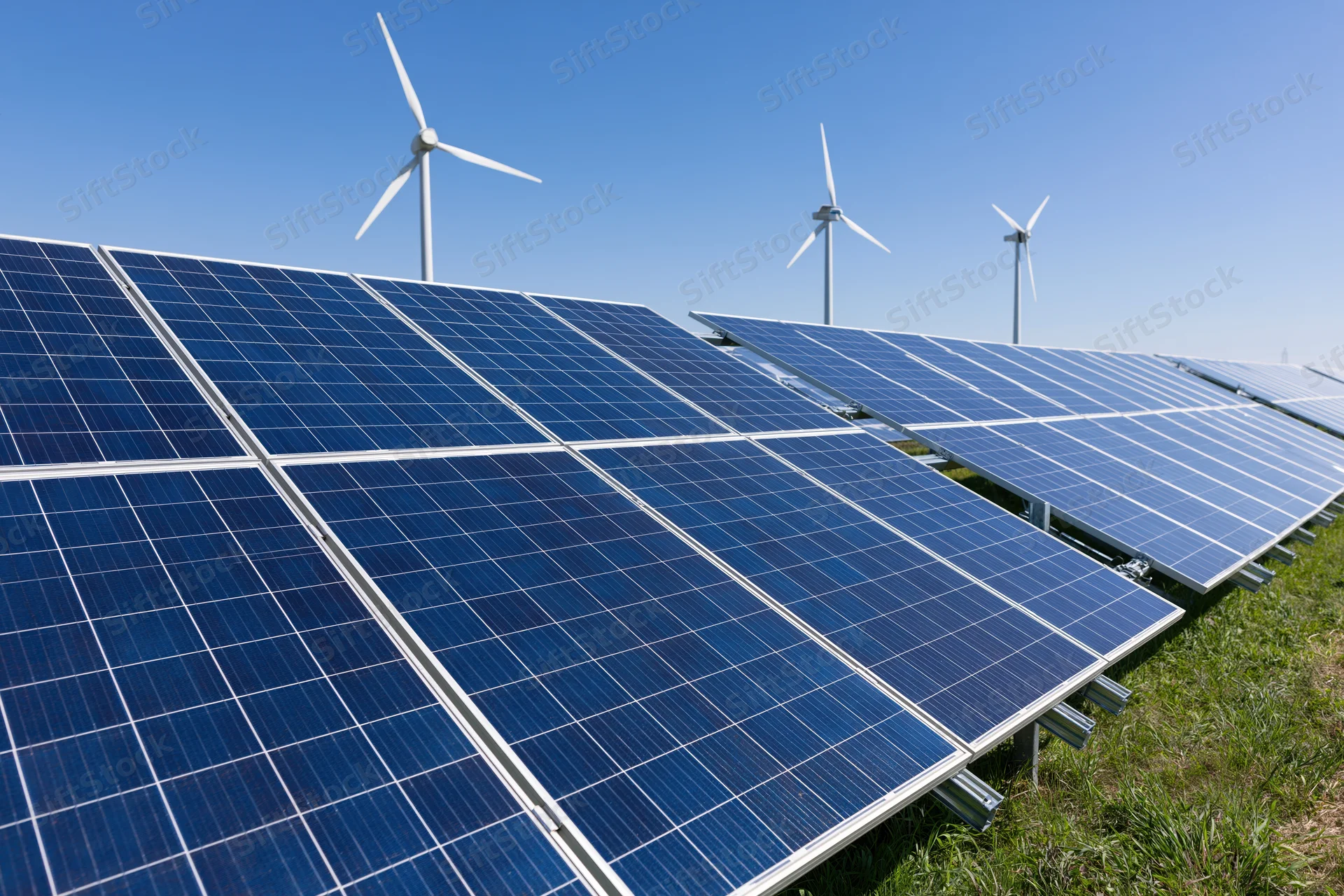 Rows of blue photovoltaic solar panels arranged in a field with wind turbines in the background under a clear blue sky