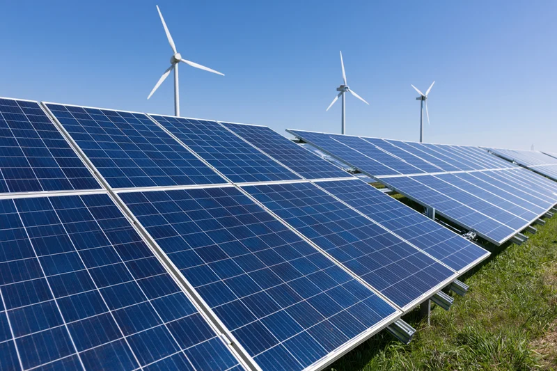 Rows of blue photovoltaic solar panels arranged in a field with wind turbines in the background under a clear blue sky