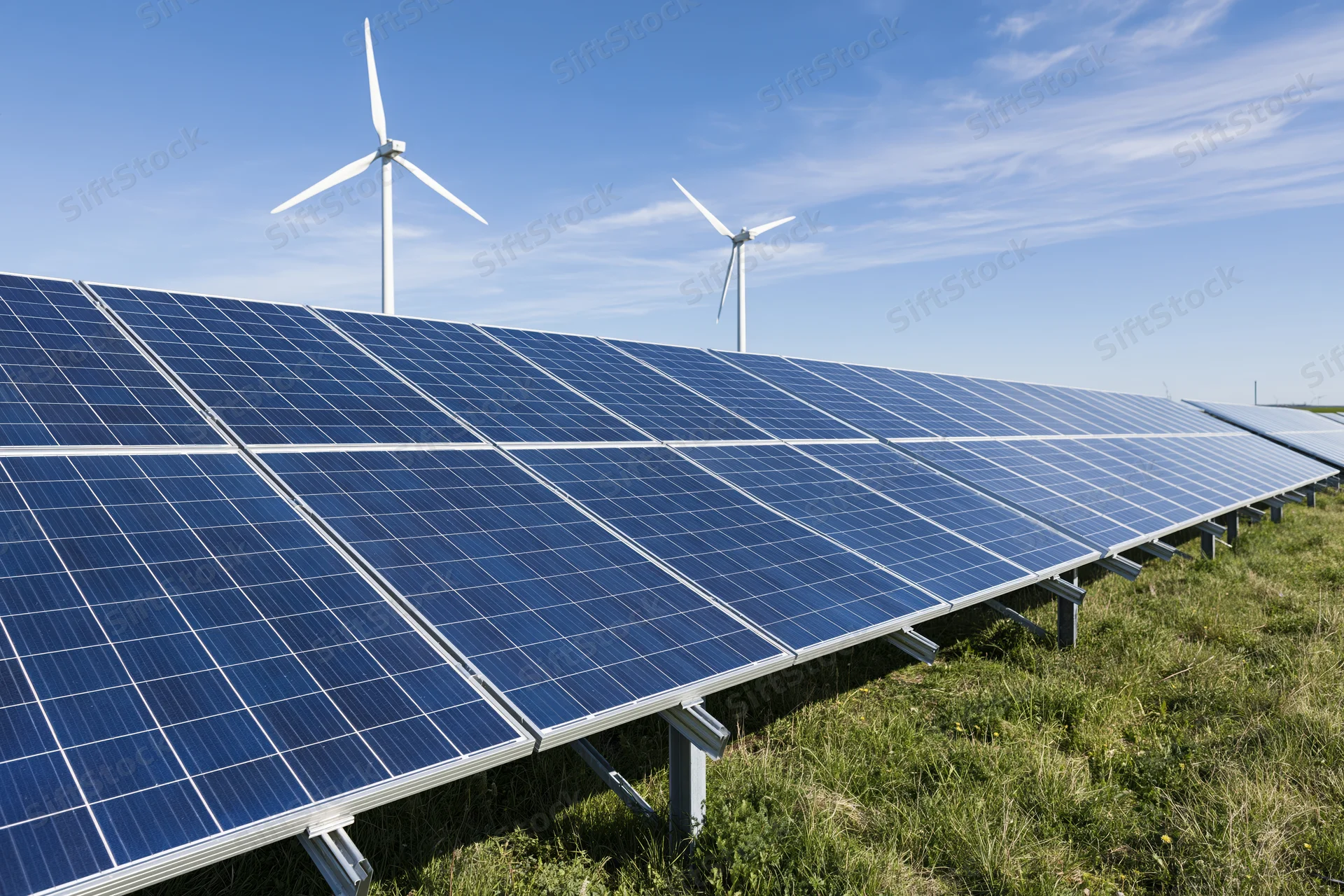 Rows of blue photovoltaic solar panels arranged in a field with wind turbines in the background under a clear blue sky