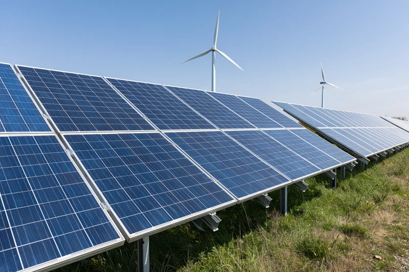 Rows of blue photovoltaic solar panels arranged in a field with wind turbines in the background under a clear blue sky